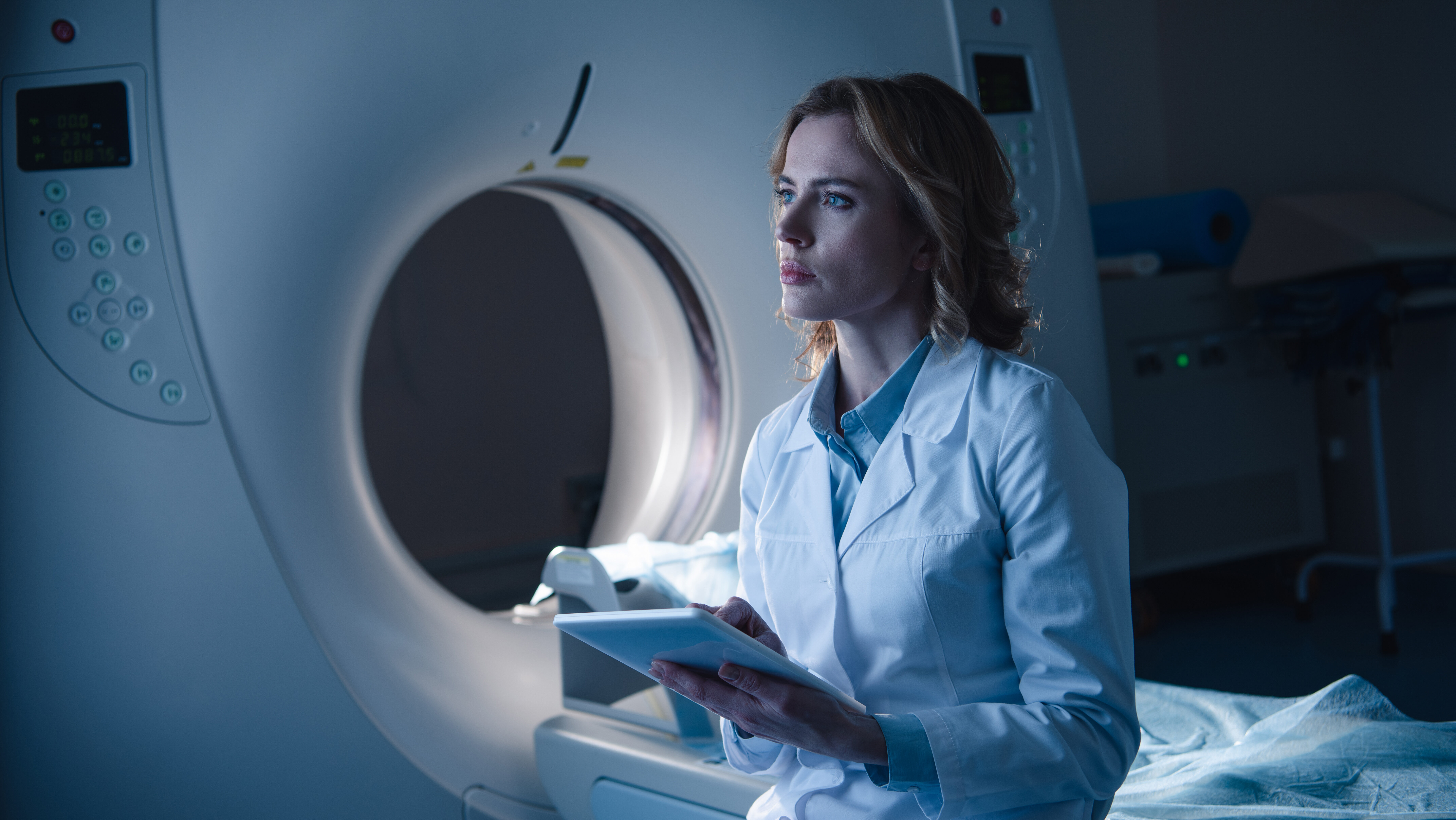serious radiologist holding digital tablet while sitting near computed tomography scanner and e1699557191357